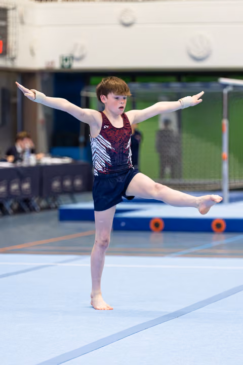 Young athlete executes a split leap with extended arms during floor exercise routine at indoor gymnastics meet