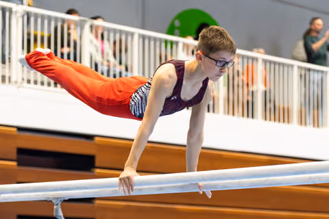 Young athlete performs horizontal balance on parallel bars while spectators watch from gallery above