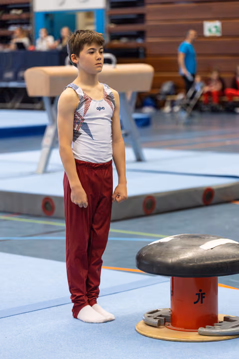 Young gymnast in white and red uniform stands attentively beside pommel horse, preparing for his routine in competition hall