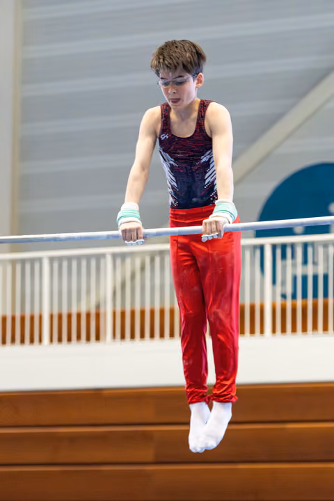 Young athlete in red pants and patterned leotard concentrates while performing on horizontal bar with white wrist guards