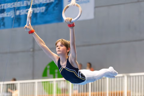 Young gymnast performs still ring hold with focused expression, arms extended during competitive routine at indoor facility