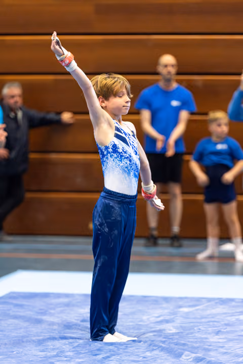 Young gymnast in blue and white leotard strikes a confident salute pose during floor exercise with spectators in background