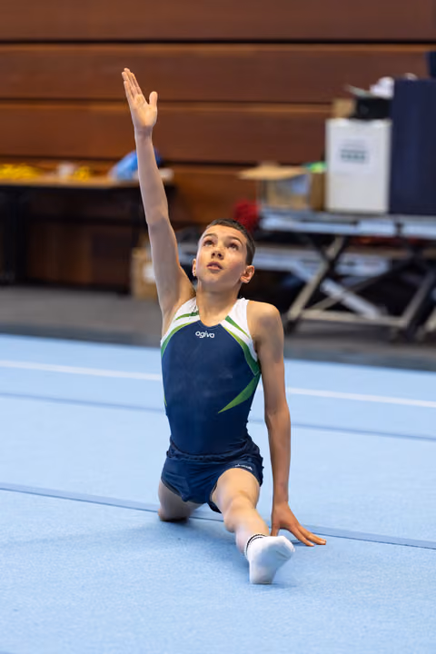 Young gymnast in navy leotard performs elegant finishing pose on floor, one arm raised gracefully while kneeling