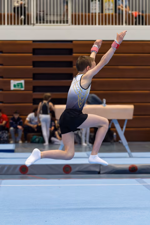 Gymnast performs split leap with raised arms during floor routine at indoor sports hall competition