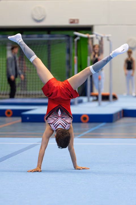 Young athlete performs a handstand with split legs on floor exercise mat, wearing red shirt and patterned shorts