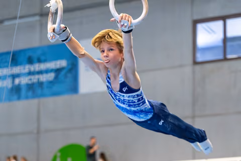 Young athlete suspended mid-air on rings, arms extended upward in perfect form during competitive routine at indoor facility