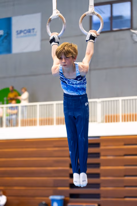 Young gymnast hangs from still rings during competition, showing strength and concentration in a sports hall