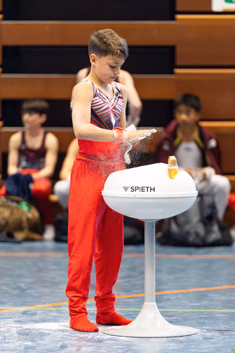 Young athlete in red outfit applying chalk to hands at Spieth apparatus before routine, with teammates seated in background