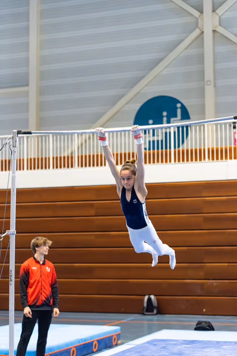 Young gymnast hanging with arms extended on high bar during training, coach observing below in indoor sports hall