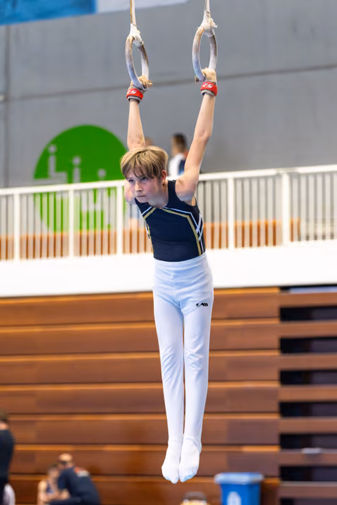 Young athlete performing rings routine with arms extended overhead, showing intense focus and concentration in gymnasium