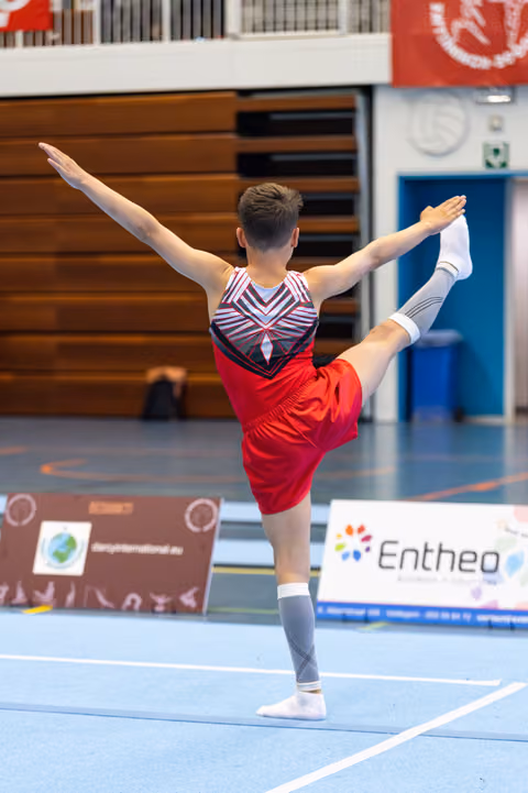 Male athlete performs graceful arabesque pose during floor routine, arms extended, wearing red and striped leotard