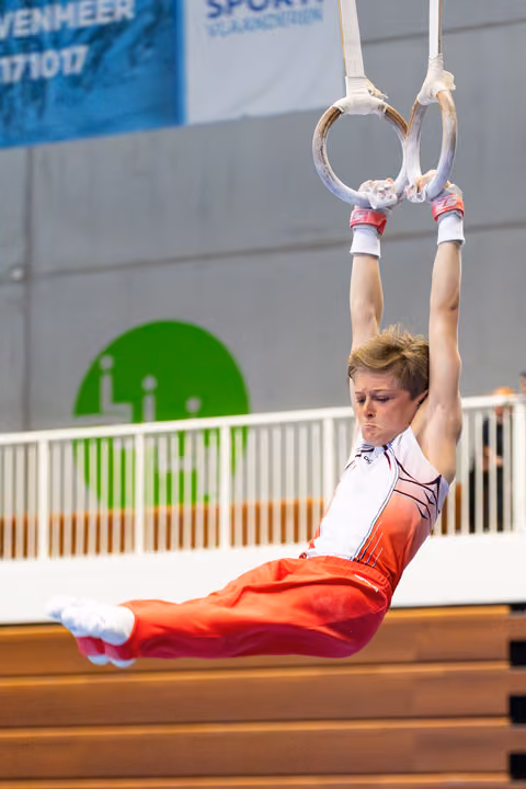 Young athlete demonstrating an L-sit position on still rings during indoor training, wearing red and white uniform