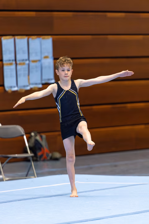 Young gymnast performs a balanced standing pose with arms extended during floor routine, displaying focus and control