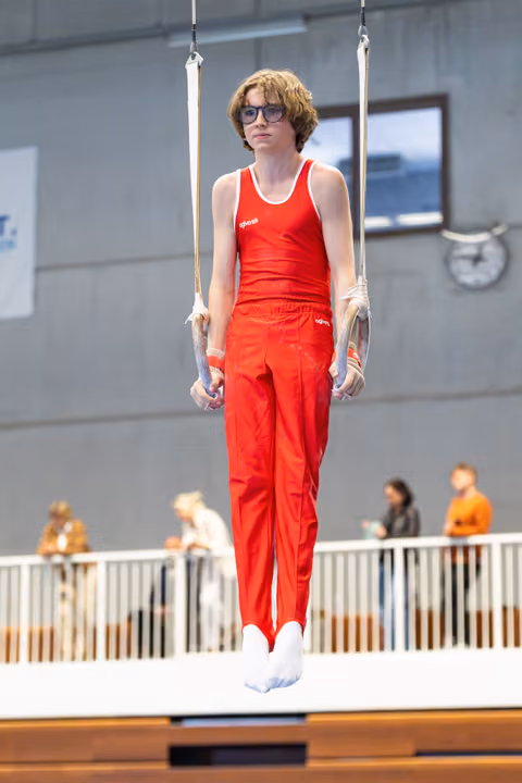 Young athlete in red uniform and glasses performing a still rings hold during indoor training session