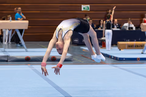 Female gymnast performing a dramatic backbend during floor routine at indoor competition, judges seated in background