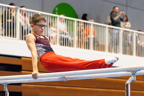 Young athlete with glasses performs parallel bars routine in intense concentration, wearing burgundy leotard and red pants