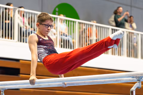 Young gymnast with glasses performs L-sit position on parallel bars while spectators watch from viewing area above