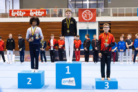 Three medal winners stand on podium positions 1-2-3, wearing their awards with proud expressions at an indoor sports venue