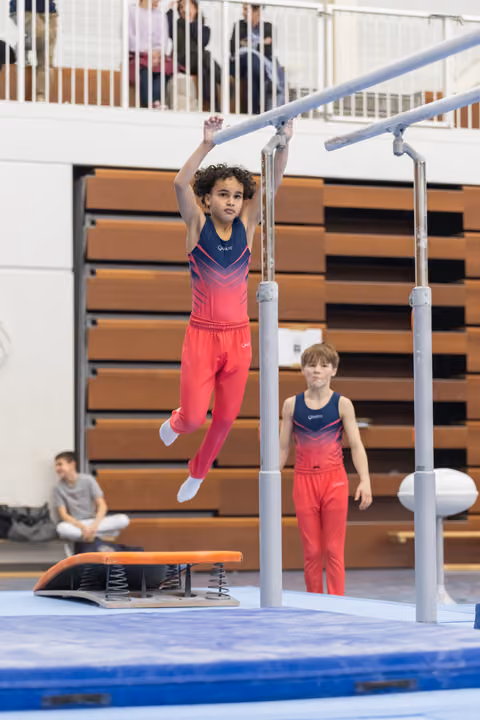 Young gymnast in pink outfit hanging from parallel bars while another child watches during training session in gymnasium