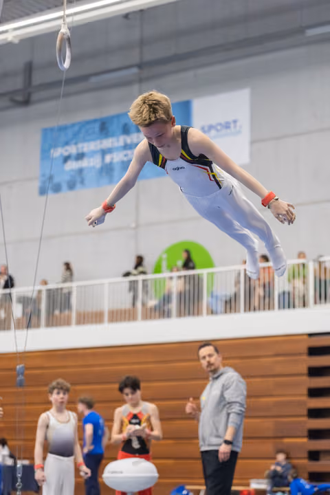 Young gymnast executes airborne trampoline dismount while coach and teammates watch from below in indoor gymnasium