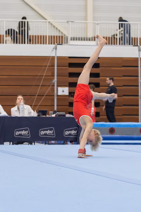 Young gymnast in red leotard performs headstand on floor mat during routine with judges table visible in background