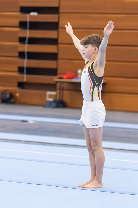Young gymnast stands with arms raised in salute pose on blue floor mat in gymnasium, displaying focused expression