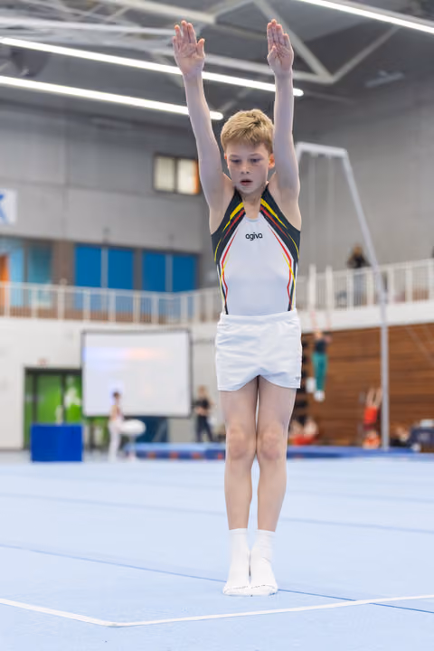 Young red-haired gymnast in white and rainbow-trimmed leotard stands with arms raised overhead in starting position