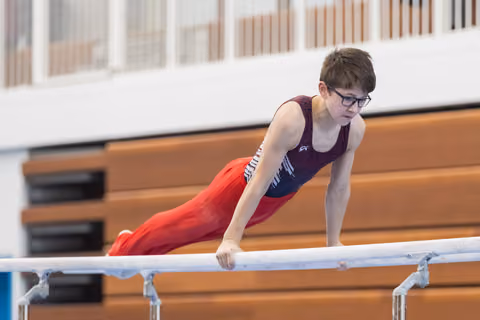 Young gymnast performs on parallel bars, body extended horizontally, wearing glasses, purple leotard and red pants
