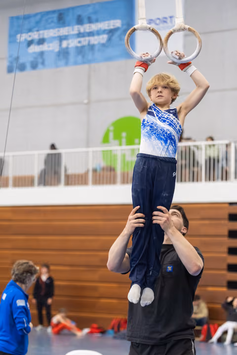 Young gymnast holds rings overhead while being lifted by coach during training session in indoor gymnasium