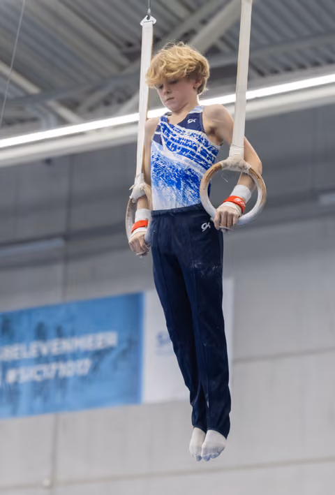 Young gymnast performing on still rings, demonstrating control and focus with bent arms in indoor training facility