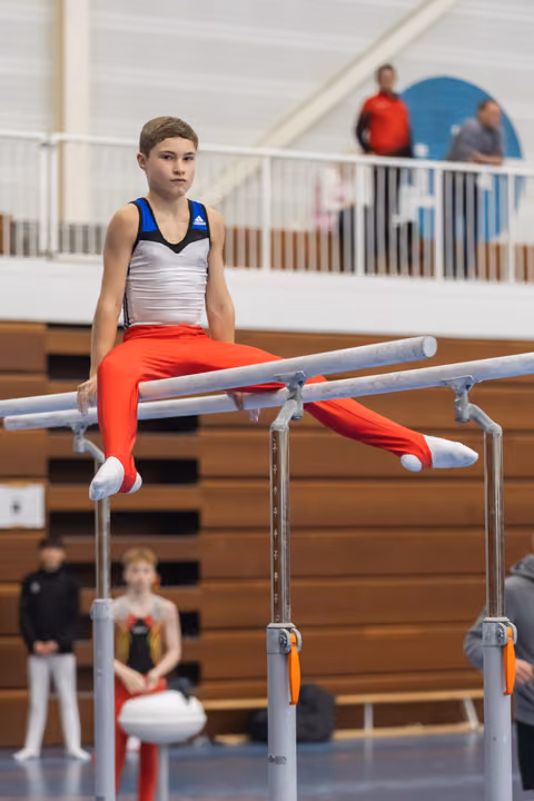 Young gymnast in red pants and white top holds a seated position on parallel bars during training session