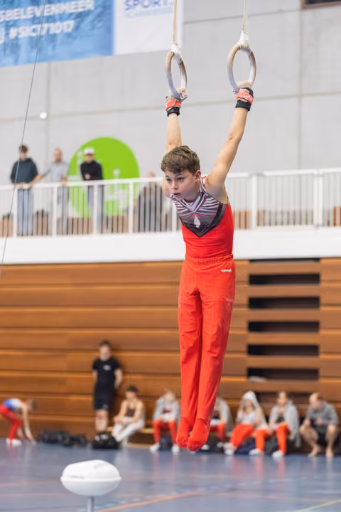 Young gymnast in red outfit hangs from rings with focused expression, arms extended overhead in indoor training facility