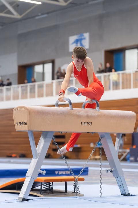 Young gymnast in red uniform concentrates intensely while performing on the pommel horse during an indoor gymnastics event