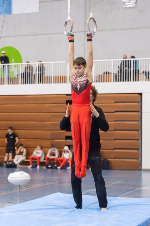 Young gymnast in red uniform performs rings routine with coach supporting from below, arms extended overhead in gymnasium