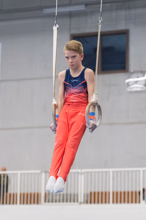 Young gymnast in coral outfit maintains focused straight-arm support position on still rings during training session