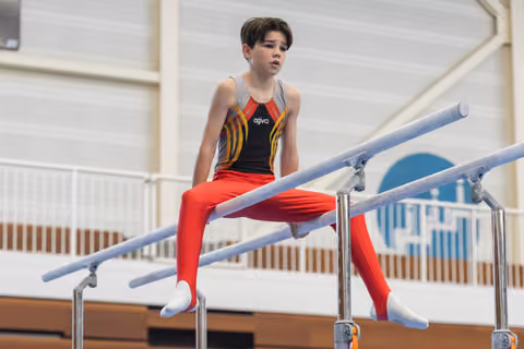 Young gymnast in colorful leotard sits on parallel bars with focused expression during training session