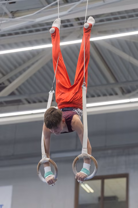 Male gymnast performs inverted handstand on rings, legs extended upward, wearing red shirt and purple shorts in training facility