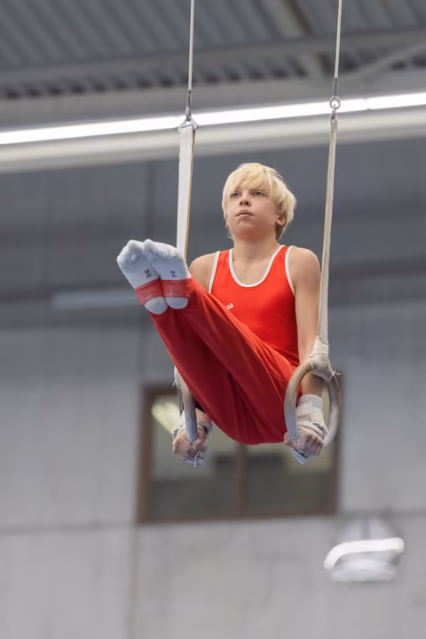 Young blonde gymnast performing on still rings in red uniform, displaying focused concentration and strength