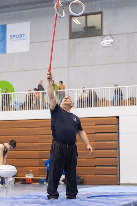 Gymnast in black uniform balances a ring on a red stick while looking up intently during an indoor performance.