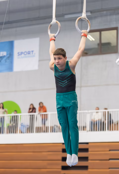 Young gymnast in teal uniform hangs from rings with focused expression during indoor gymnastics event