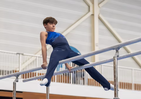 Young gymnast in blue leotard executes a horizontal hold on parallel bars, displaying strength and concentration in training facility