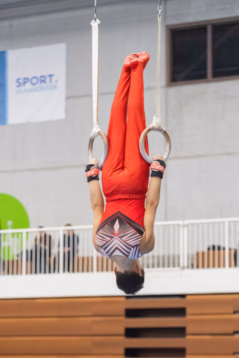 Gymnast performs inverted hang on rings with extended body, wearing red outfit during indoor training session
