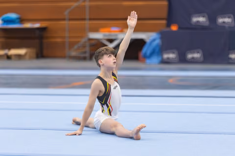 Young gymnast completes floor routine with raised arm, sitting in splits position on blue mat in training facility