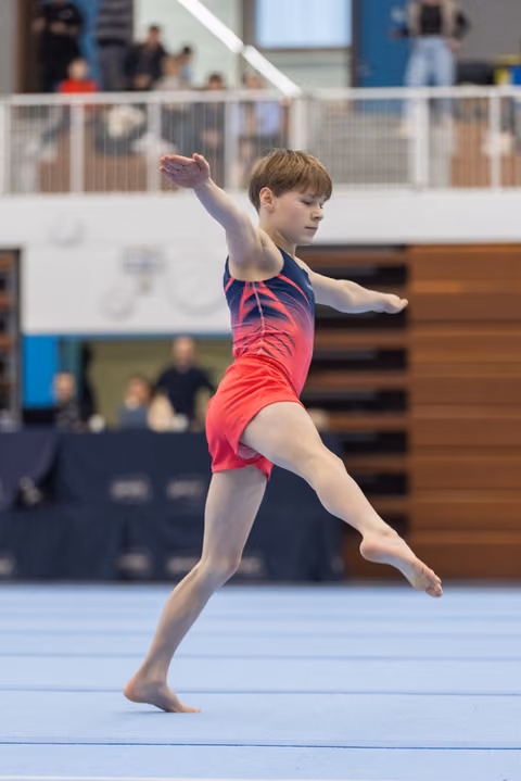 Young gymnast performs an elegant dance element during floor routine, arms extended gracefully in pink and navy leotard