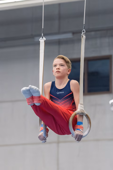 Young gymnast performs rings routine with determined expression, wearing navy and red leotard in training facility