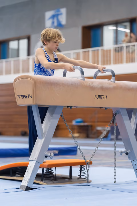 Young blonde gymnast gripping pommel horse handles with intense concentration during practice at indoor gymnasium facility