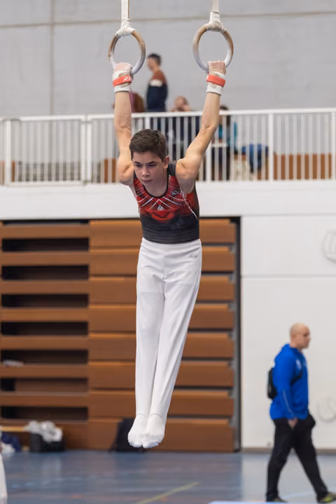 Young male gymnast hangs from still rings with arms extended, showing intense concentration during routine in gymnasium