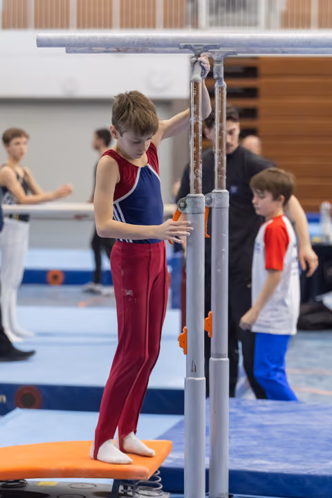 Young gymnast in maroon and blue leotard adjusts parallel bars height while other athletes practice in background