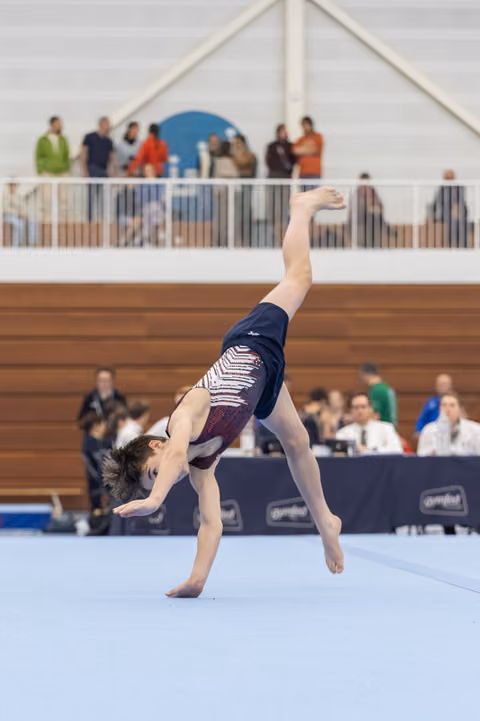 Gymnast performs dramatic backward arch on floor exercise, demonstrating flexibility before judges and spectators