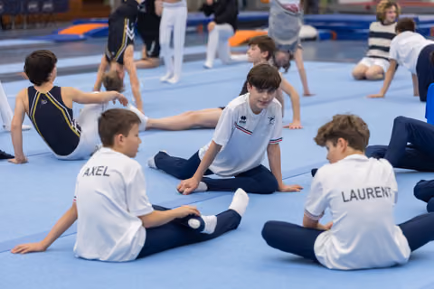 Young gymnasts in white polo shirts perform floor stretches during warmup, with names like 'AXEL' and 'LAURENT' on their backs
